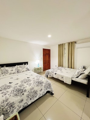 A hotel room with two beds, a wooden door, a small table with a lamp, and a window with curtains. - Casa Miel: Sweet Getaway in Paradise (Puerto Jiménez)