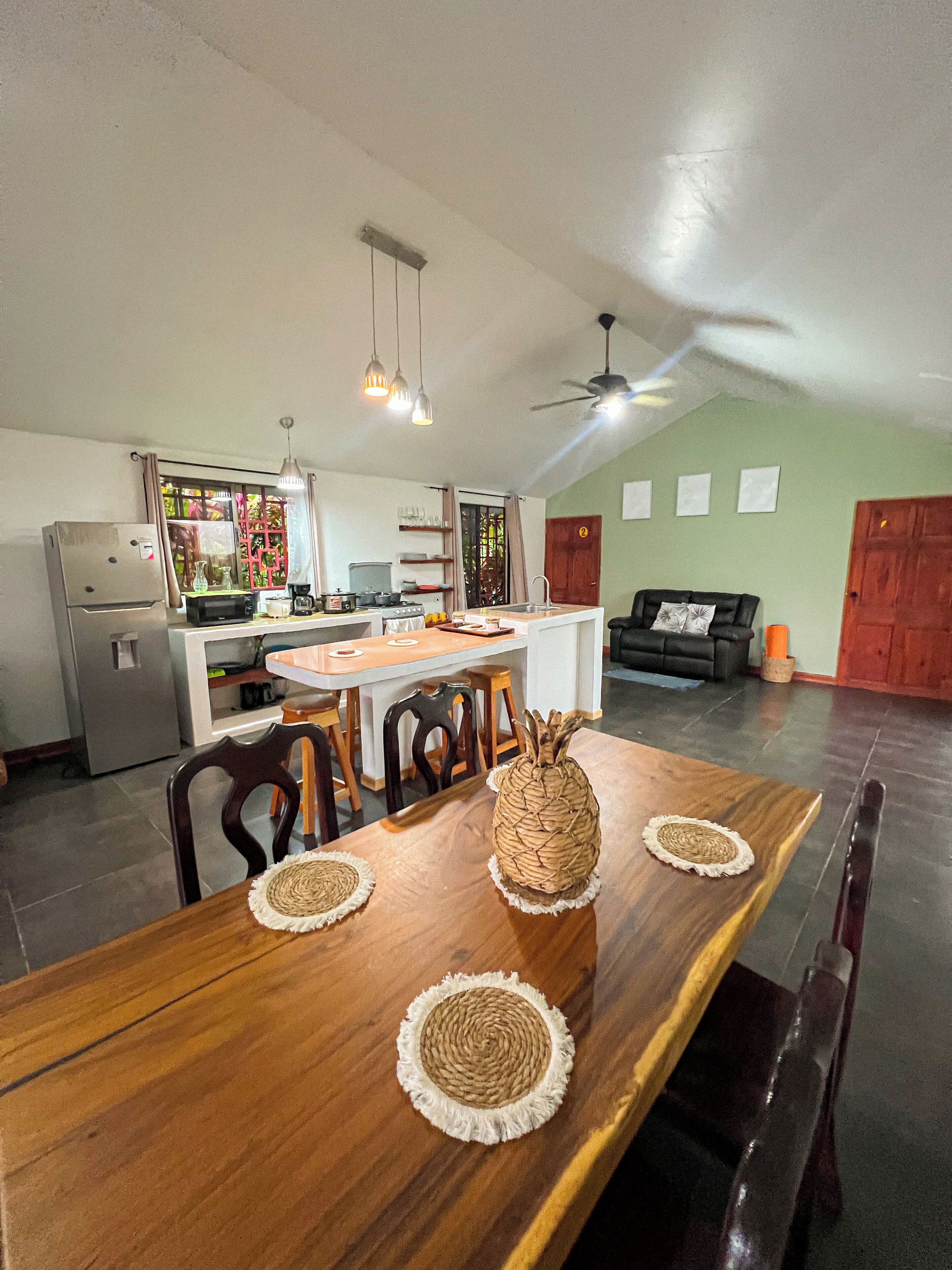 A spacious living area with a wooden dining table, chairs, and a woven centerpiece. A kitchen with modern appliances and a dining area with a sofa and a coffee table are visible.