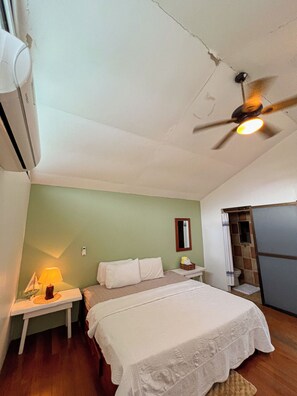A bedroom with a bed, nightstand, lamp, ceiling fan, and a small bathroom with a shower. - Casa Miel: Sweet Getaway in Paradise (Puerto Jiménez)