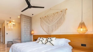 Une chambre spacieuse avec un grand lit, un ventilateur de plafond, une décoration murale en macramé et un luminaire suspendu.