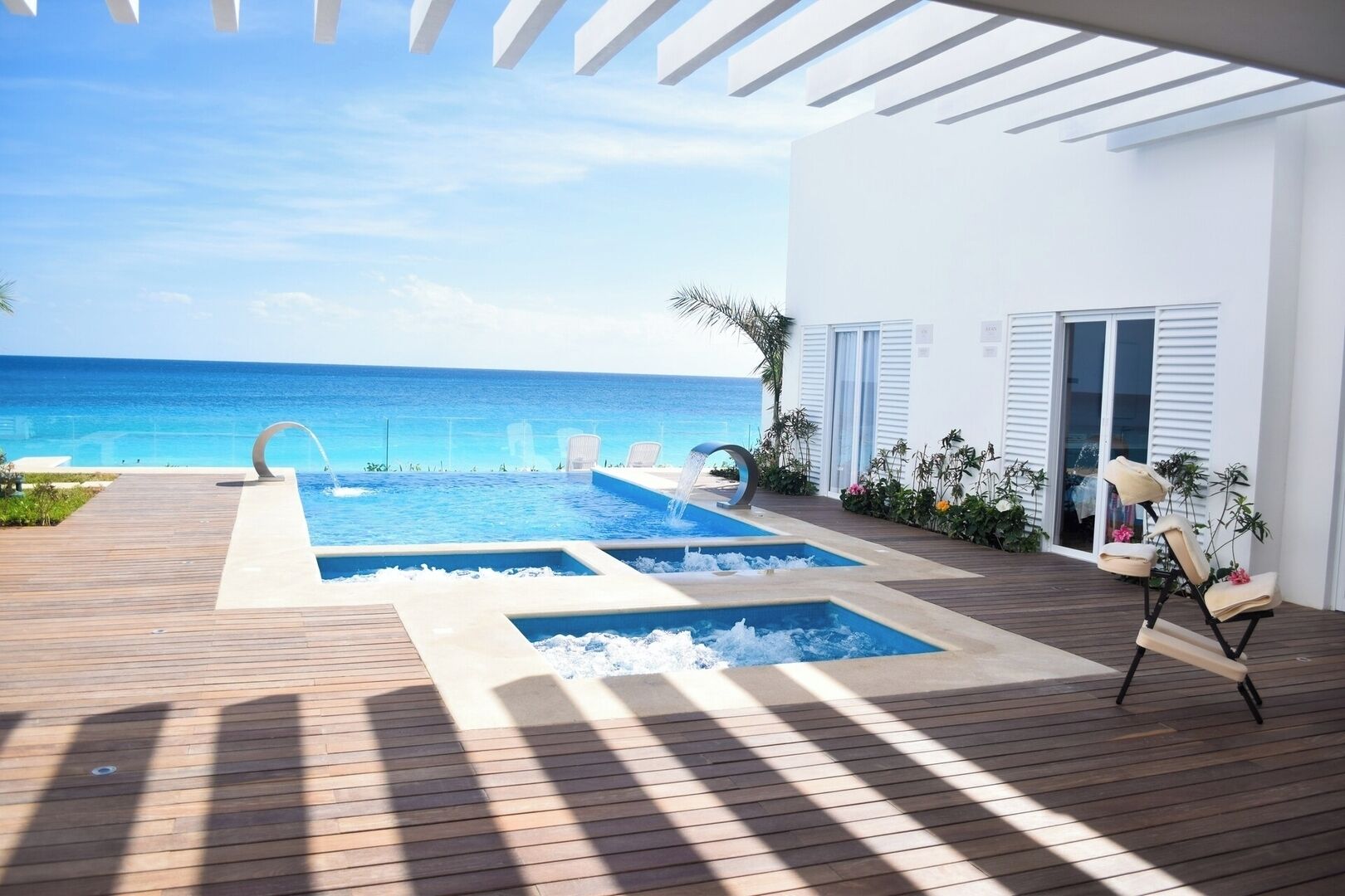 Zona de piscina moderna al aire libre con vista al océano, cubierta de madera y sillones de descanso.