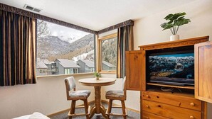 Quarto de hotel com uma grande janela, um armário de madeira com uma televisão, uma mesa redonda com duas cadeiras e uma cama.