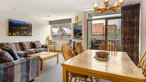 Uma sala de estar aconchegante com um sofá, uma mesa de jantar com cadeiras, uma televisão e uma janela grande com vista para as casas.