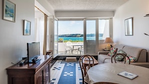 A room with a television, a dining table, a sofa, and a view of the sea.