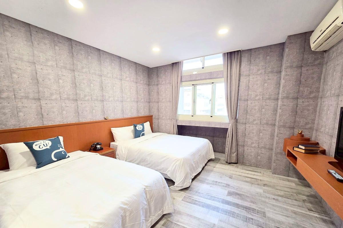 Habitación de hotel con dos camas, cabecero de madera, ventana con cortinas, televisor sobre un mueble de madera y aire acondicionado instalado en la pared.