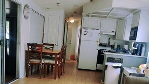 Eine kleine Küche mit Kühlschrank, Herd und Spüle. Es gibt eine Essecke mit Tisch und Stühlen. Der Boden ist Holzboden, die Wände sind weiß.