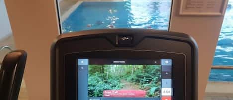 Máquina de ejercicio Matrix con pantalla mostrando un video, ubicada en una sala con vista a una piscina cubierta.
