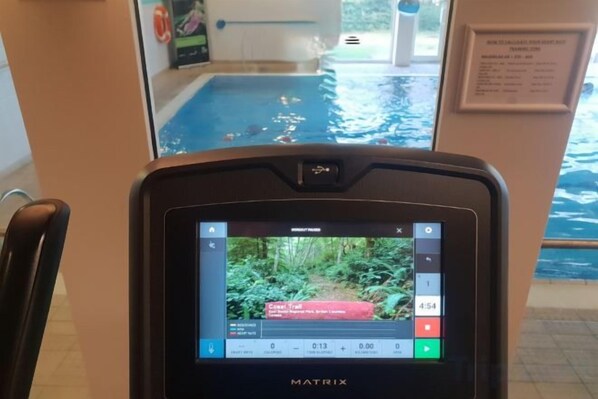 A Matrix exercise machine with a screen displaying a video, located in a room with a view of an indoor swimming pool.