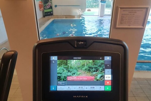 A Matrix exercise machine with a screen displaying a video, a pool view, and a sign with text. - Holiday Inn COLCHESTER by IHG (Eight Ash Green)