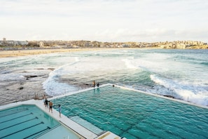 Une piscine en bord de mer, avec une eau d’un bleu cristallin, entourée d’une plage et avec une vue sur la ville en arrière-plan.