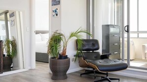 Ett modernt vardagsrum med ett stort fönster, en svart läderfåtölj, en golvlampa i rotting och en säng i bakgrunden.
