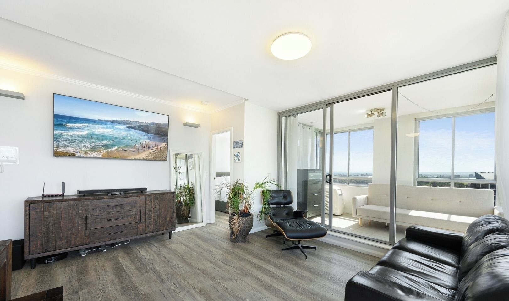 Un soggiorno moderno con una grande TV a schermo piatto, un divano in pelle, una porta vetrata che dà su un balcone e un mobile in legno.