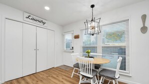 Une pièce comprenant une table à manger, des chaises, un lustre, un panneau indiquant l’emplacement de la buanderie et une fenêtre avec des stores.