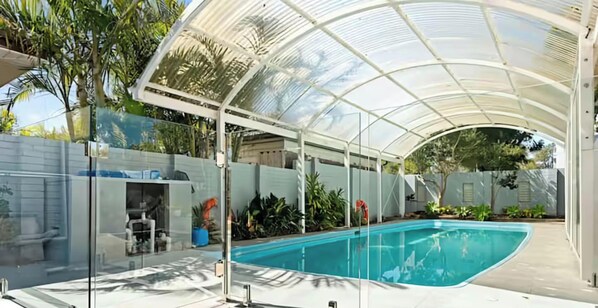 En swimmingpool under et glastag med udsigt til haven og en hvid væg.
