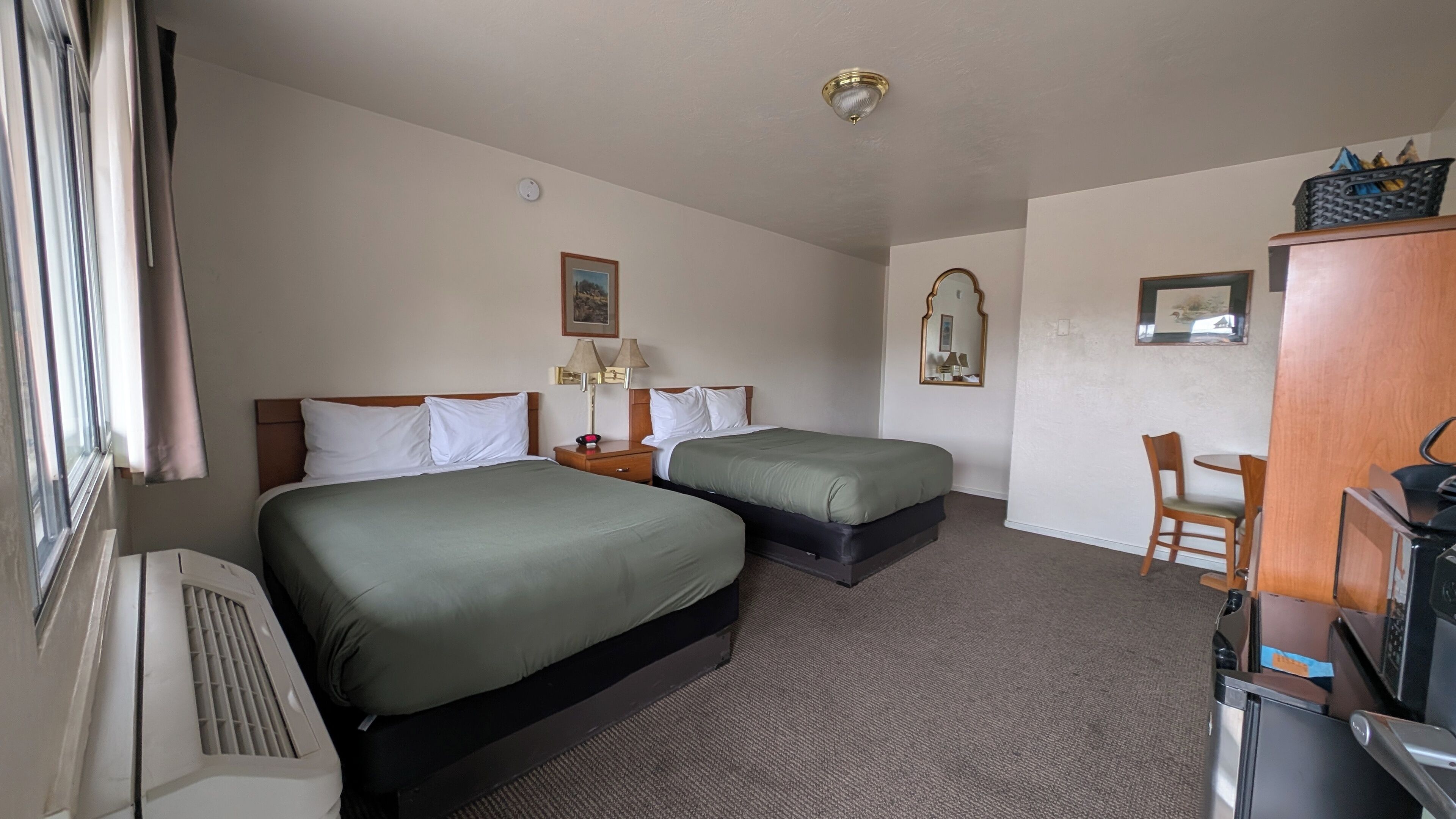 A hotel room with two beds, a desk, a chair, a heater, a mirror, and a ceiling light.