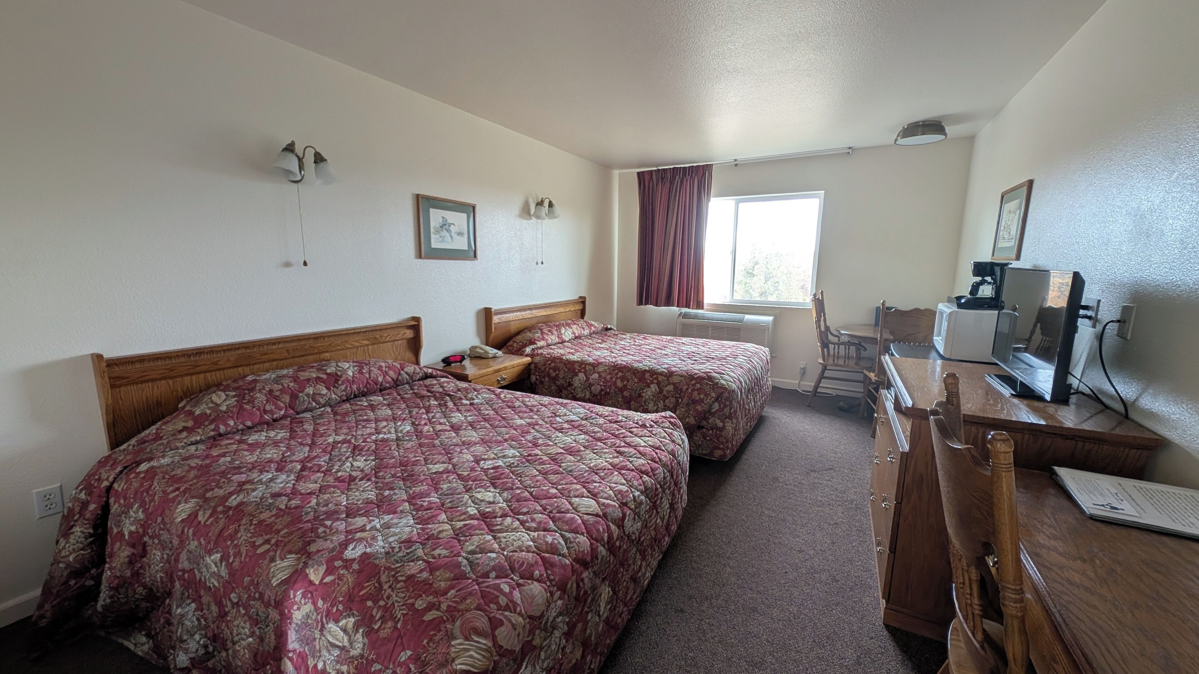 Habitación de hotel con dos camas, un escritorio con computadora, una silla y un ventanal con cortinas.