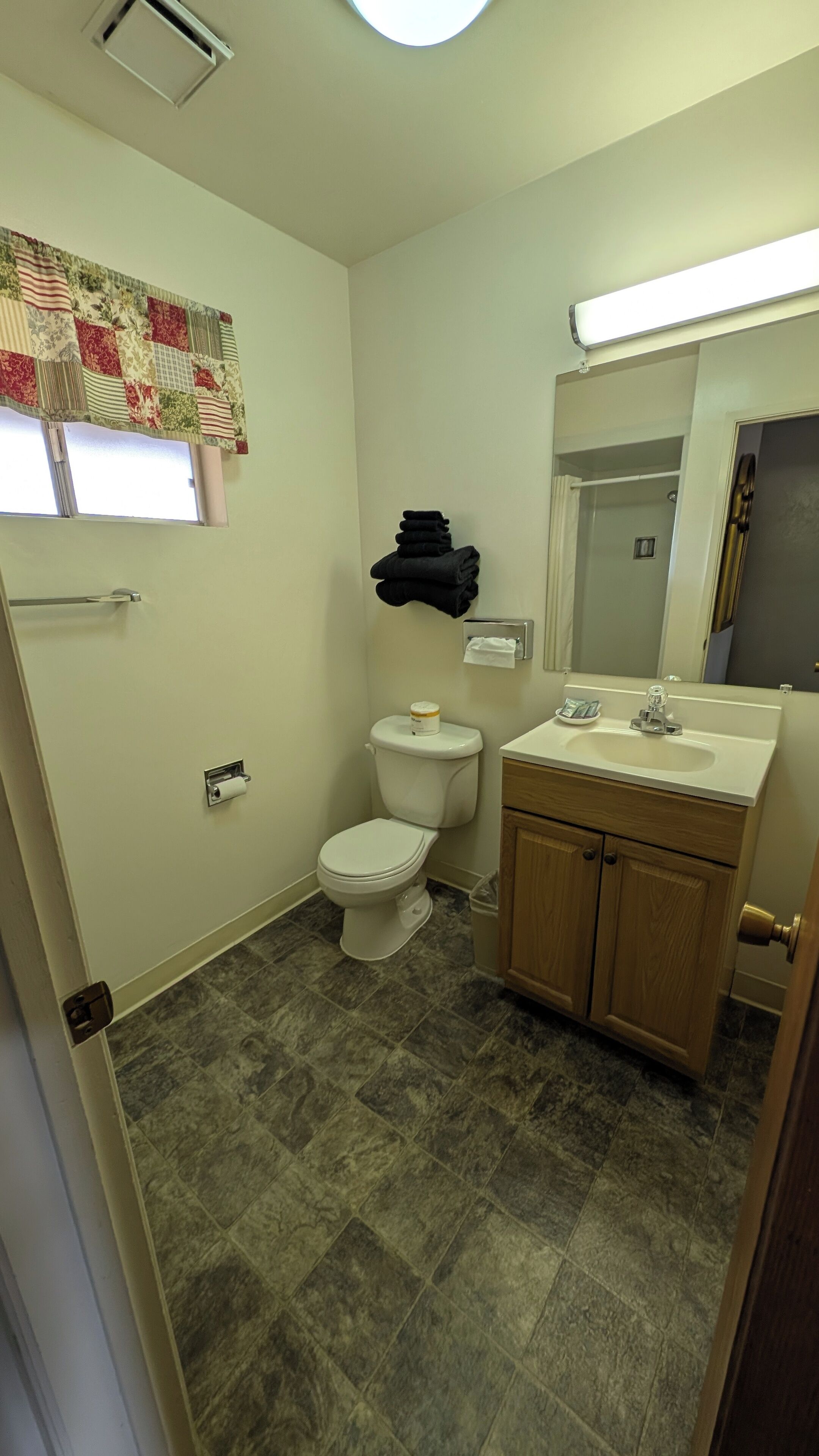 Et badeværelse med toilet, vask, spejl og et vindue med blomstret gardin.