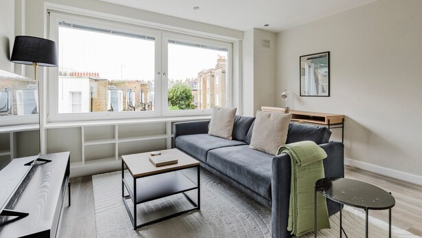 Ein modernes Wohnzimmer mit grauem Sofa, einem kleinen Holztisch, einer Lampe und Blick auf die Stadt durch große Fenster.