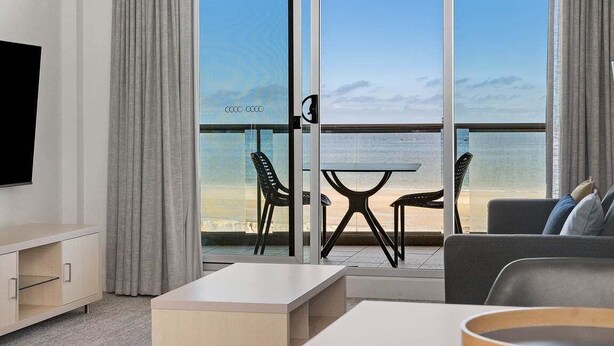 Ein modernes Hotelzimmer mit einer großen Schiebeglasdoor zum Balkon mit Blick auf den Strand. Ein Flachbildfernseher ist an der Wand montiert, es gibt ein Sofa und einen Couchtisch.
