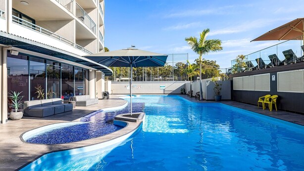 Una piscina de diseño curvo, rodeada de sillas de descanso y sombrillas, y un edificio con balcones y ventanas amplias.