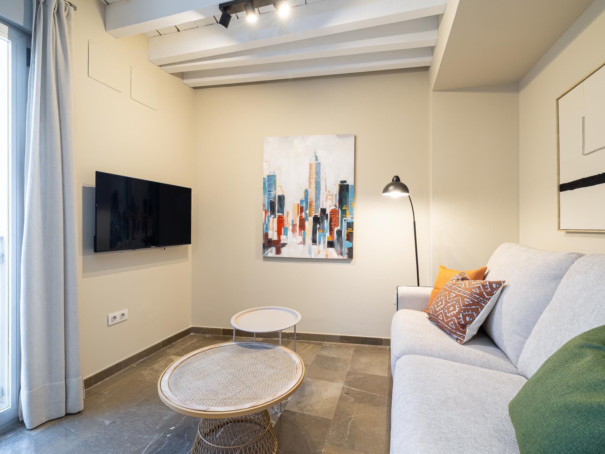 En moderne stue med et fladskærms TV, et bybillede som maleri, et rundt fletbord, en grå sofa med mønstrede puder og en gulvlampe.