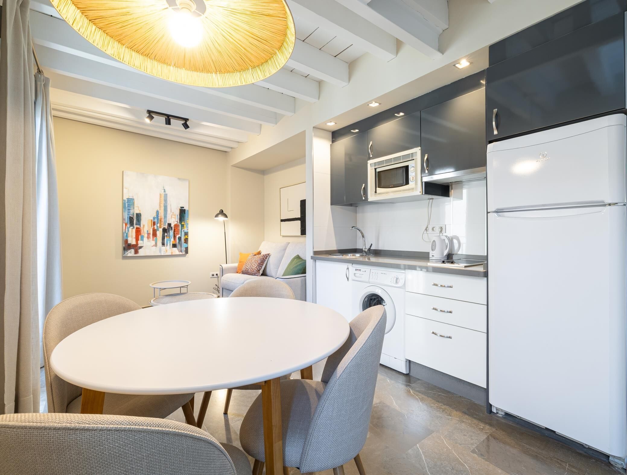 A modern kitchen with white appliances, a dining area with a round table and chairs, and a cityscape painting on the wall.