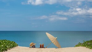 Escena de playa con una duna de arena, un sombrero de paja, una bebida y una sombrilla.