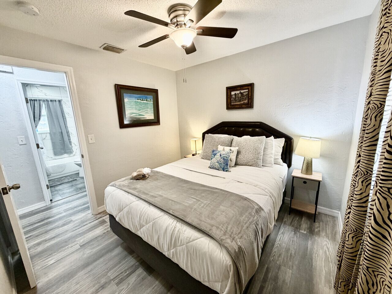A hotel room with a bed, ceiling fan, two bedside lamps, and a door leading to a bathroom.