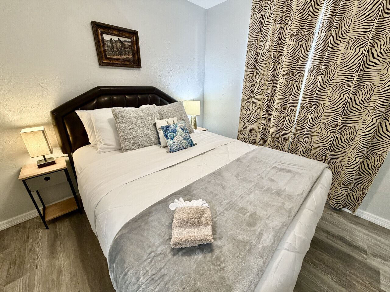 Un lit bien fait, avec une couette grise et une serviette pliée, une table de chevet avec une lampe, et un rideau à motifs zébrés.