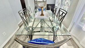 Een eethoek met een glazen tafel, omringd door stoelen, een decoratieve metalen plank met objecten, en een uitzicht door dunne gordijnen.