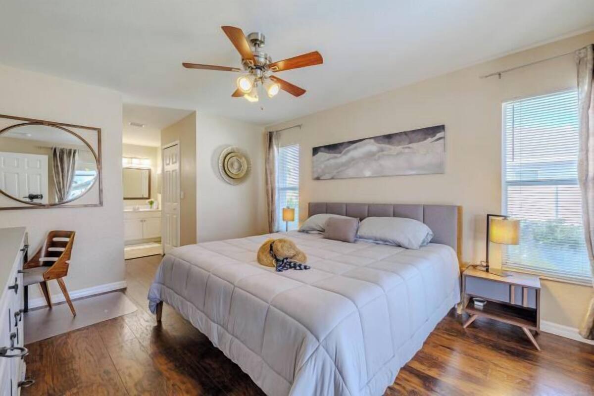 Une chambre à coucher comprenant un lit, un ventilateur de plafond, une table de chevet, une lampe, une fenêtre avec des stores, une porte et un miroir.