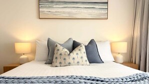 Un lit bien fait, recouvert d’une couverture en grosse maille, avec deux lampes de chevet et un ensemble de coussins sur le thème de la plage.