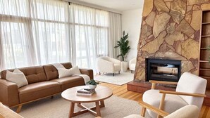 Un salon moderne avec un canapé en cuir marron, une table basse ronde en bois et deux fauteuils blancs. On aperçoit une cheminée en pierre au fond.