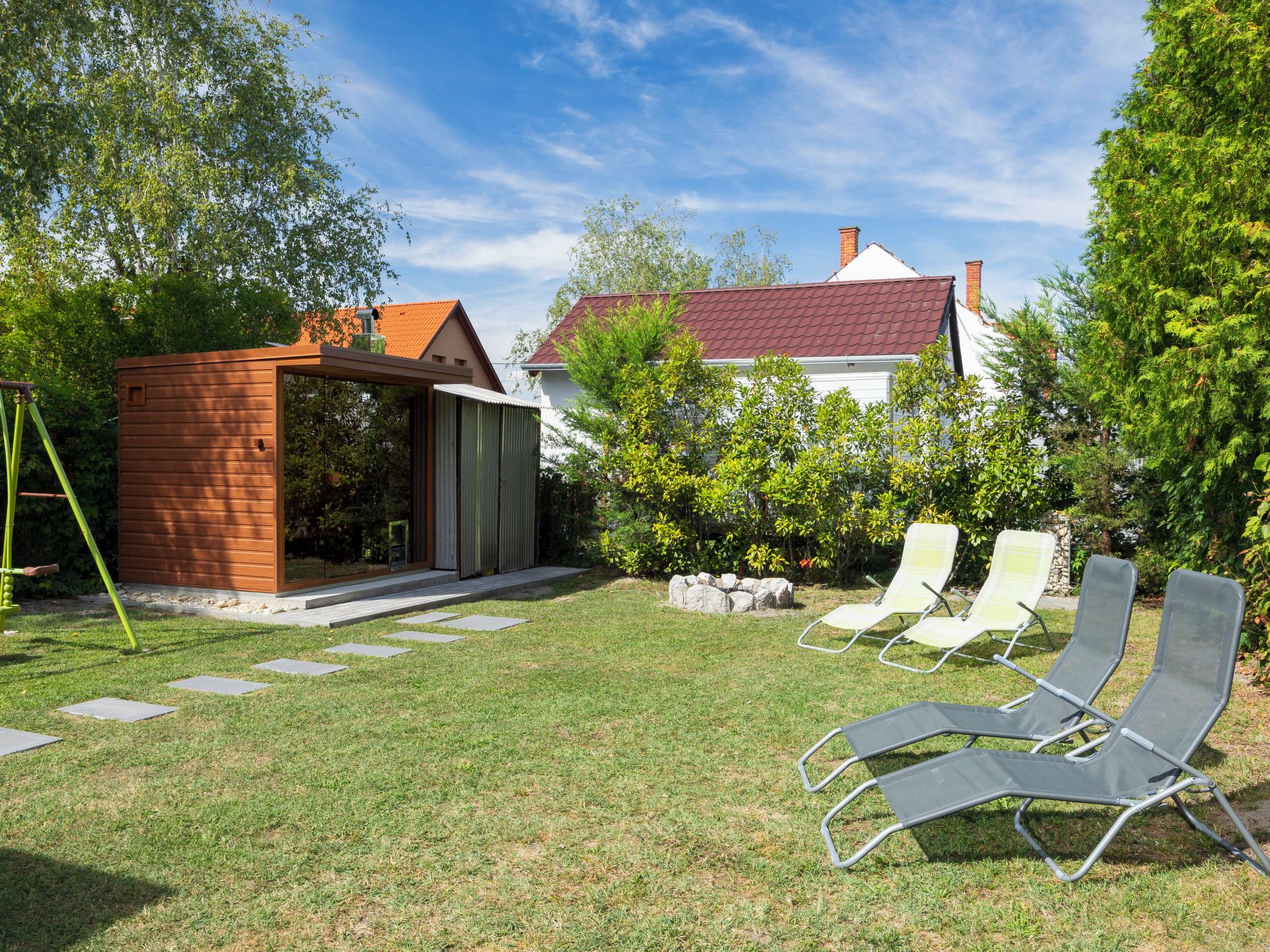 A backyard with a small wooden structure, a white house with a red roof, and two lounge chairs.