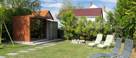 A backyard with a small wooden structure, a white house with a red roof, and two lounge chairs.