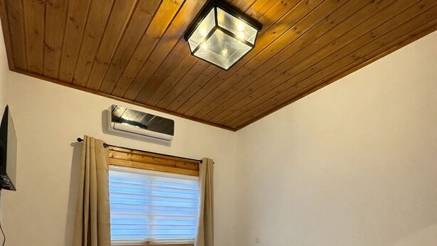 A bedroom with a wooden ceiling, a bed with white bedding, a window with curtains, and an air conditioning unit. - Beautiful 9-bedroom villa with AC in serene Gshur (Gshur)
