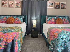 Ein Zimmer mit zwei Betten, jeweils mit einer farbenfrohen, gemusterten Tagesdecke, einem Nachttisch mit Lampe und einer kleinen Schublade.