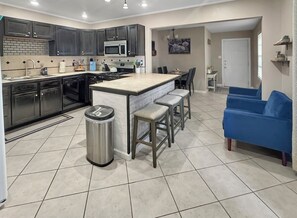 A modern kitchen with dark cabinets, a central island, and a blue sofa in the living area. - 3BD Retreat| 5 Mins to Downtown! (Abilene)