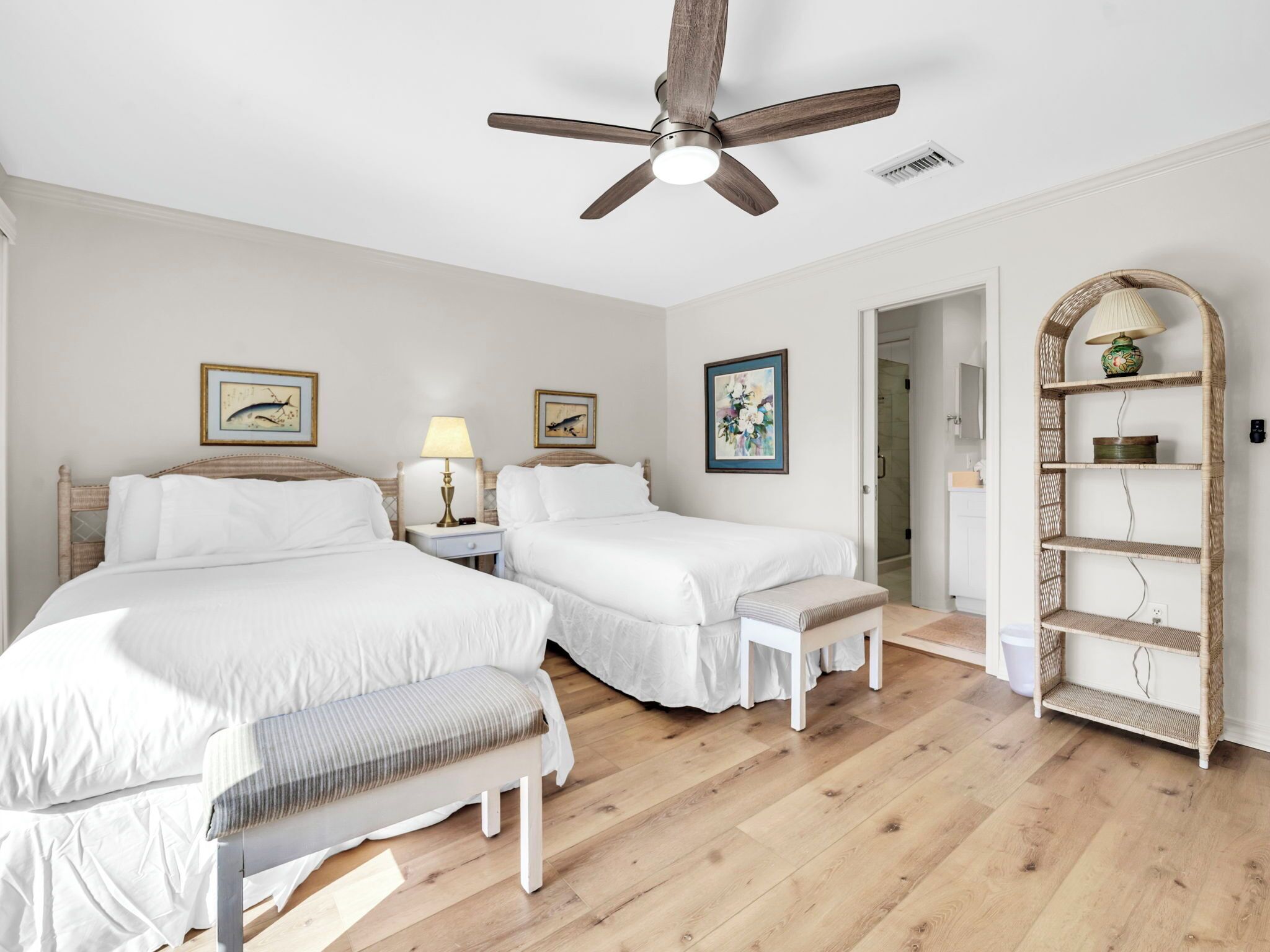 A bedroom with two beds, a ceiling fan, a bedside table with a lamp, and a wicker shelf.