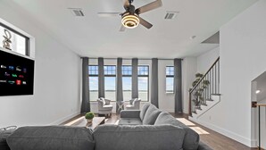 A modern living room with a grey sofa, a ceiling fan, large windows, and a staircase.