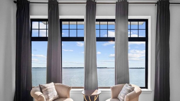 A room with two beige armchairs, a small round table, and a view of the sea through large windows with dark curtains.