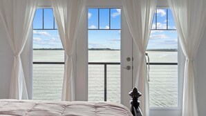 A bedroom with a large window overlooking a body of water, curtains, and a bed.