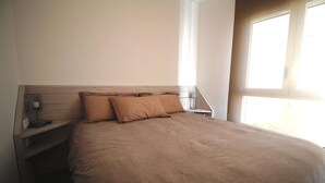 A neatly made bed with a headboard, a bedside table with a lamp, and a window providing natural light. - CALA FONDA Apartment in Santa Margarita Roses (ROSES)