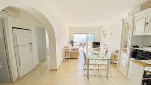 A spacious living area with a glass dining table, a sofa, and a TV. There is a kitchen with a refrigerator and cabinets. - CENTRAL Apartment Roses near the beach with sea view (Roses)