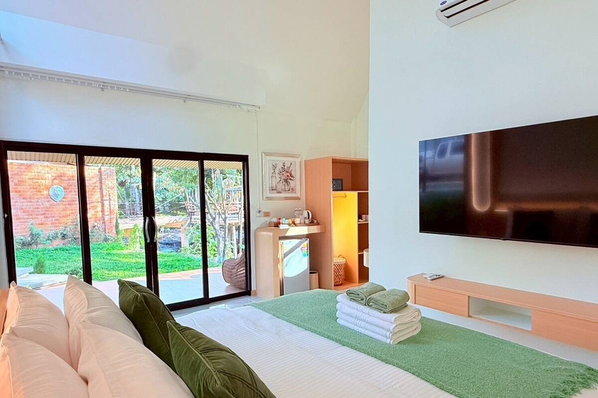 A modern hotel room with a large bed, a flat-screen TV, a green rug, and a view of a garden through a sliding door.