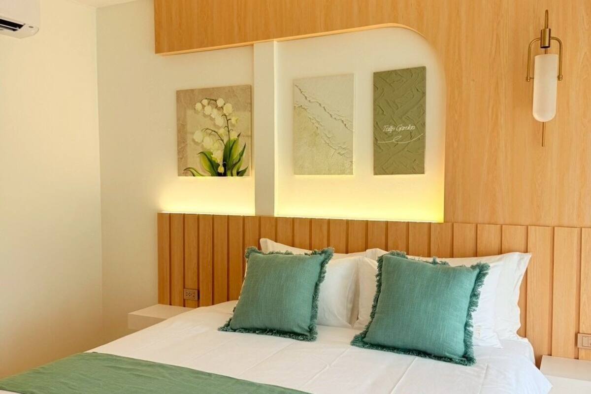 A modern bedroom with a wooden headboard, a bed with white linens and green pillows, and three framed artworks on the wall.