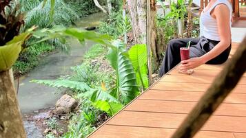 A person sitting on a wooden deck overlooking a river, with a thatched roof structure and lush greenery in the background.
