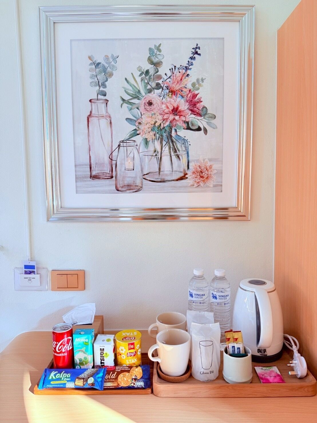 A framed floral artwork on a wall, a coffee maker, two water bottles, a coffee cup, a mug, a glass, a tray with snacks, and a card reader.