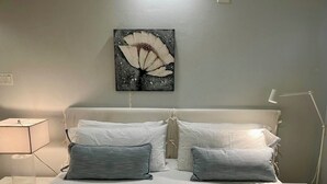 Een keurig opgemaakt bed met witte beddengoed en twee grijze kussens, een bedlampje en een aan de muur bevestigd kunstwerk van een bloem.