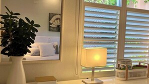 Een kamer met een bed, een nachtkastje met een lamp, een potplant en een raam met zonwering.